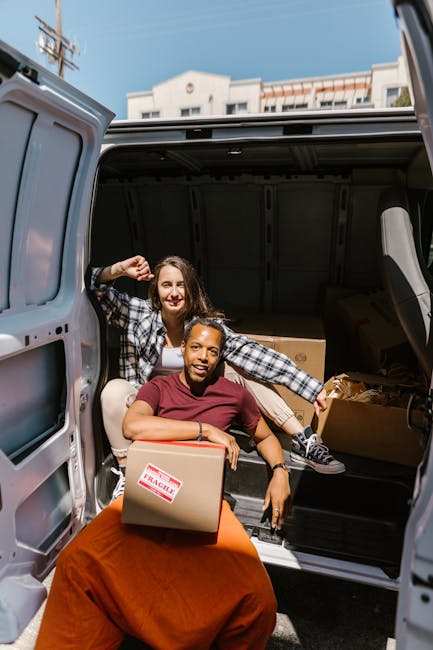 Two women are seen during the home relocation process, working together to load or unload boxes from the rear of a grey van parked outdoors under a clear sky. One woman, wearing a checkered shirt and jeans, is seated inside the van, smiling and holding a cardboard box. The other woman, dressed in a maroon top and orange skirt, is sitting on the ground next to the open van door, also smiling, while holding a labeled cardboard box. The van's interior is visible, showing more moving boxes and packing materials, including cardboard and plastic-wrapped items. In the background, a building with antennas and utilities is partially visible, indicating an urban environment. The scene captures elements of packing, furniture transport, and the loading process typical of local removals, consistent with services offered by Enfield Removals, specialists in house and furniture moving.
