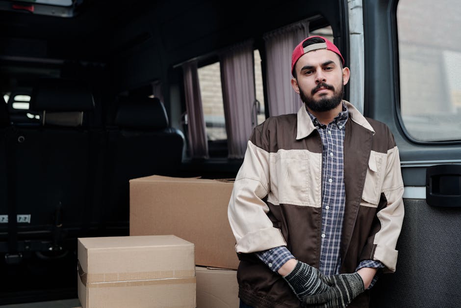 Two women are seen during the home relocation process, working together to load or unload boxes from the rear of a grey van parked outdoors under a clear sky. One woman, wearing a checkered shirt and jeans, is seated inside the van, smiling and holding a cardboard box. The other woman, dressed in a maroon top and orange skirt, is sitting on the ground next to the open van door, also smiling, while holding a labeled cardboard box. The van's interior is visible, showing more moving boxes and packing materials, including cardboard and plastic-wrapped items. In the background, a building with antennas and utilities is partially visible, indicating an urban environment. The scene captures elements of packing, furniture transport, and the loading process typical of local removals, consistent with services offered by Enfield Removals, specialists in house and furniture moving.