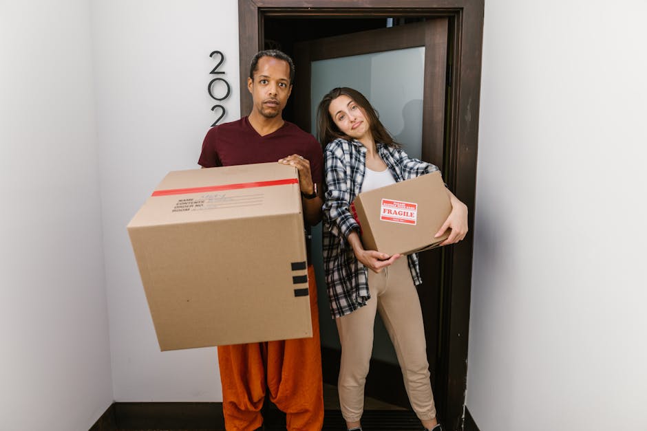 A man and a woman stand inside a building at a doorway, each holding a cardboard box used for packing during a home relocation. The man on the left wears an orange pair of trousers and a burgundy t-shirt, holding a large cardboard box with black stripes and red tape on top. The woman on the right wears beige trousers and a blue, black, and white checkered shirt, holding a smaller cardboard box labeled Fragile with red and white stickers. The doorway behind them leads outside and is framed with dark wood, with partly visible house numbers on the wall to the left. The interior has plain white walls and a dark floor, illuminated by natural or ceiling lighting, reflecting the packing and moving process involved in house furniture transport, packing, and loading for a large house removal, a service occasionally provided by Enfield Removals.